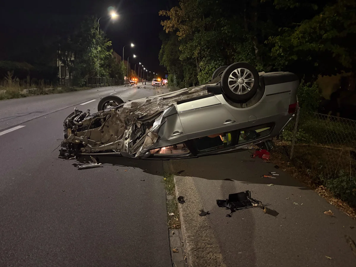 Ein Auto liegt bei Nacht auf dem Dach auf einer Straße, umgeben von Trümmern. Straßenlaternen beleuchten die Szene leicht.
