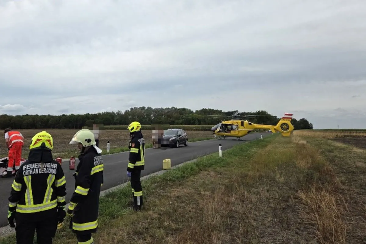 Feuerwehrleute in Uniform stehen auf einer Landstraße, während ein gelber Rettungshubschrauber im Hintergrund gelandet ist. Auf der Straße befinden sich auch ein Auto und weitere Einsatzkräfte.