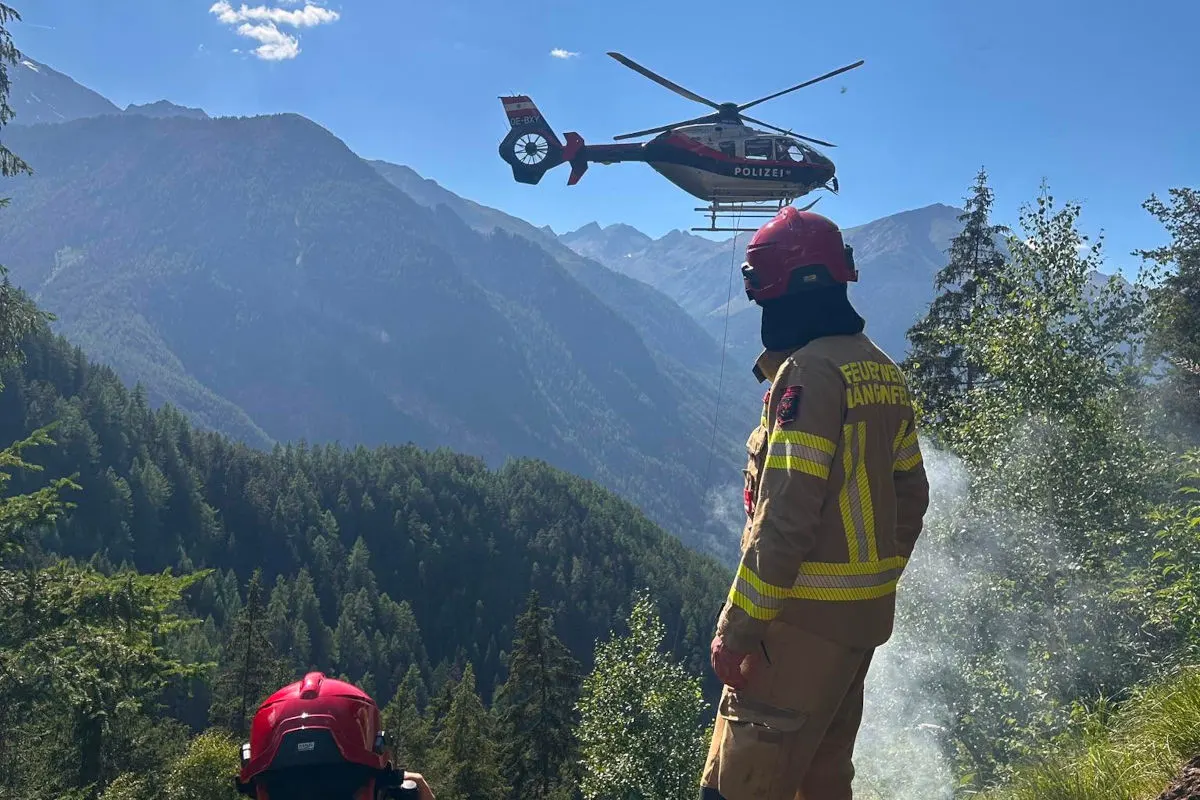 Ein Feuerwehrmann steht auf einem Berg und beobachtet einen in der Luft schwebenden Polizeihubschrauber. Um ihn herum sind Bäume und Berggipfel zu sehen.