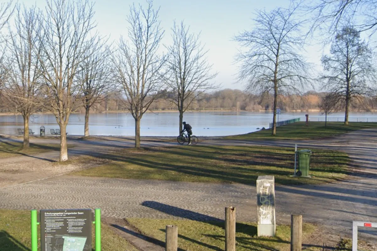 Ein Radfahrer fährt auf einem Weg neben Bäumen, im Hintergrund ein ruhiger See bei klarem Wetter.