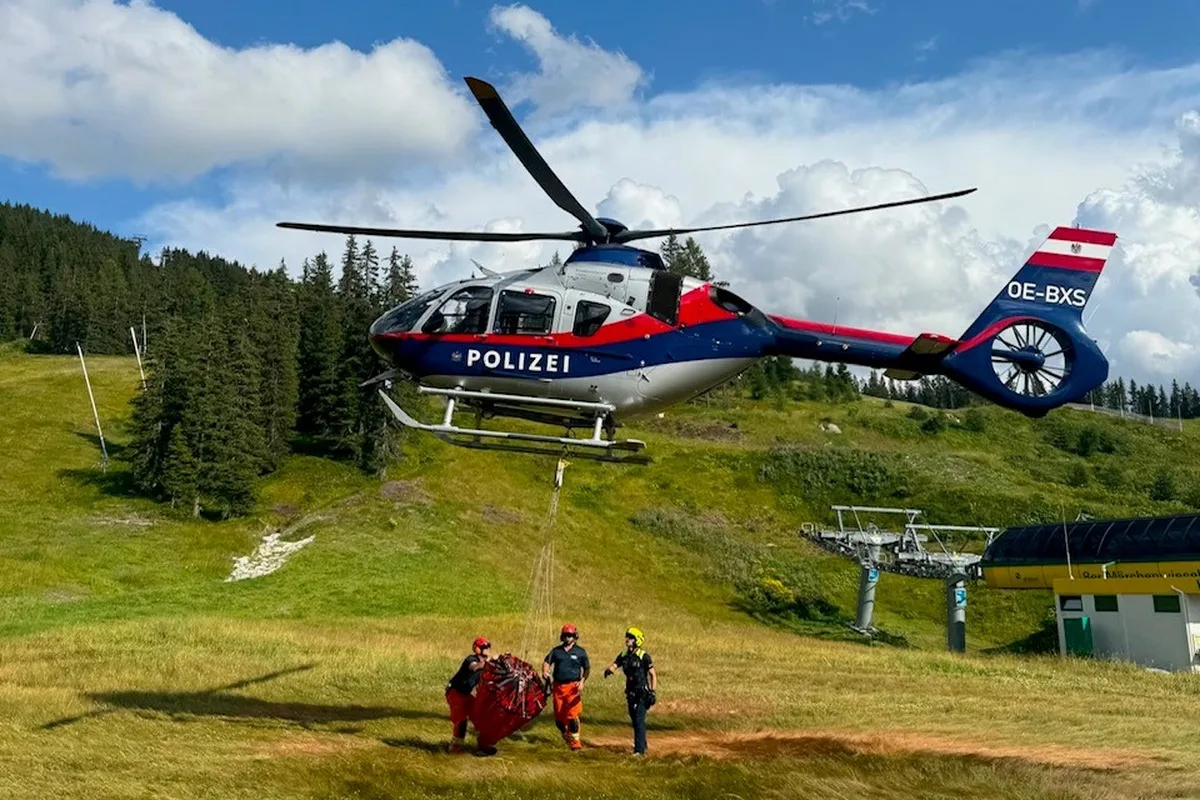 Ein Polizeihubschrauber in blau und rot schwebt über einer grünen Wiese in den Alpen. Am Boden stehen drei Personen in Rettungskleidung.