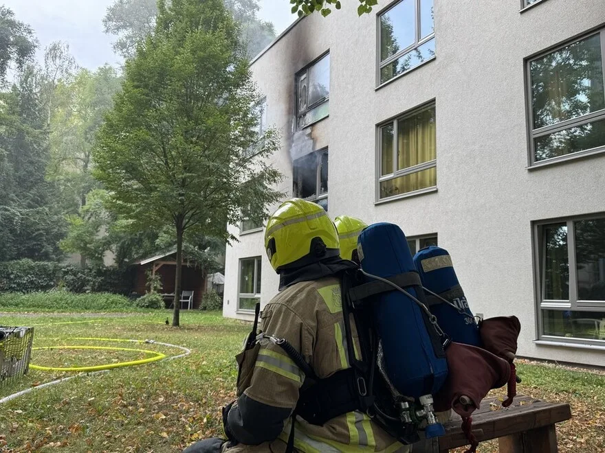 Zwei Feuerwehrleute mit Atemschutz sitzen vor einem mehrstöckigen Gebäude, aus dem Rauch dringt. Löschschläuche sind auf dem Boden verteilt.