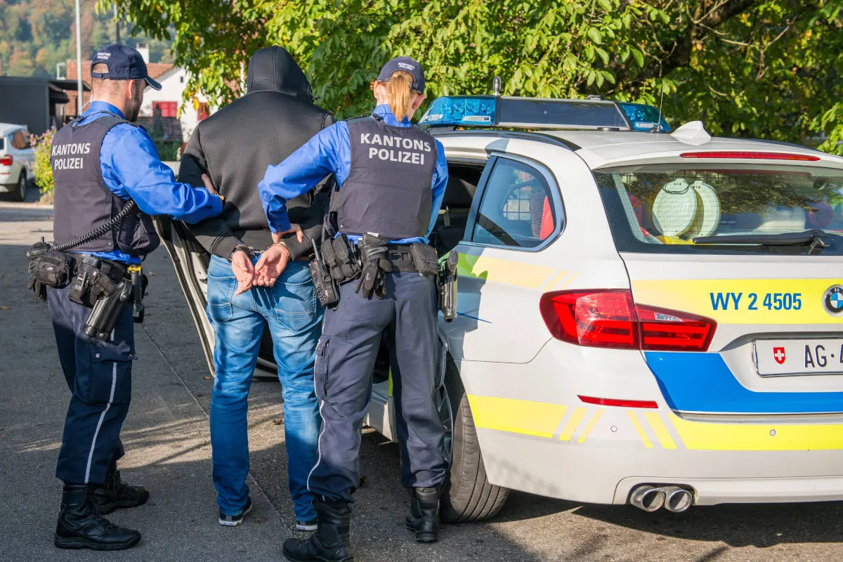 Zwei uniformierte Polizisten führen eine Person mit überkreuzten Händen hinter dem Rücken zu einem Polizeiauto mit geöffneter Tür.