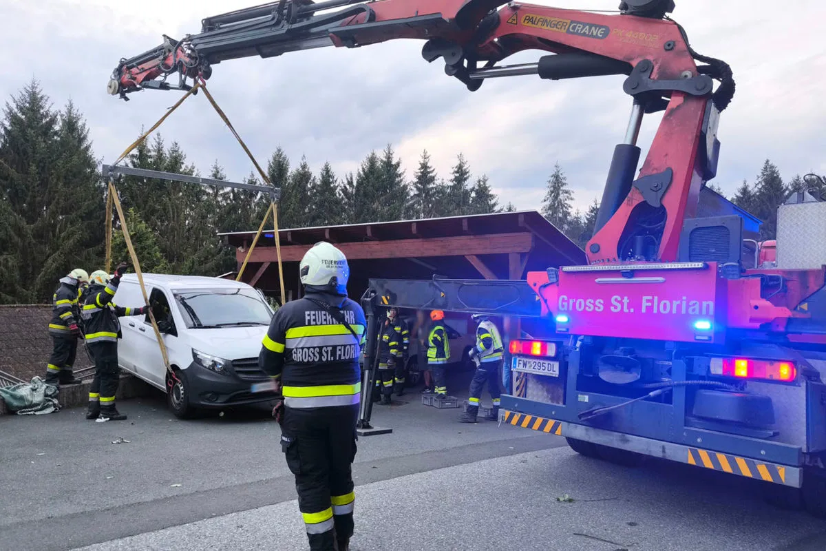 Mehrere Feuerwehrmänner sind damit beschäftigt, einen weißen Transporter mit einem Kran anzuheben. Im Hintergrund ist ein Carport zu sehen.