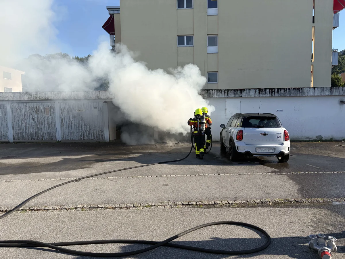 Zwei Feuerwehrleute in gelben Helmen bekämpfen ein Feuer in einer stark rauchenden Garage. Ein weißer Mini Cooper ist daneben geparkt.