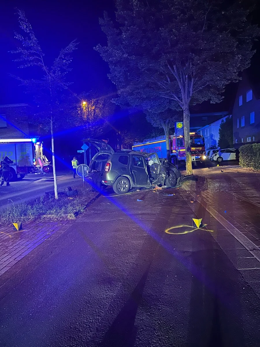 Ein Auto ist bei Nacht an einen Baum gefahren, umgeben von Einsatzfahrzeugen und Markierungen auf der Straße.