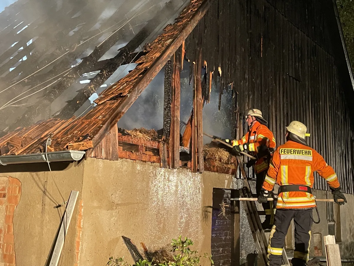 Zwei Feuerwehrleute in orange-gelben Uniformen löschen einen Dachstuhlbrand bei Nacht. Dicker Rauch ist zu sehen.