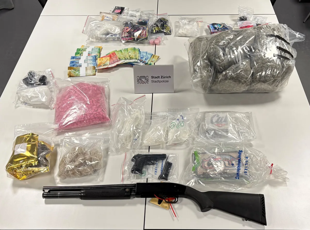 Auf einem weißen Tisch liegen in Tütchen verpackte Drogen, Geld in Schweizer Franken und zwei Waffen, darunter eine Schrotflinte.