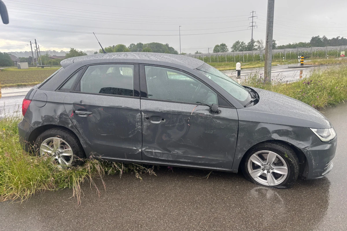 Ein grauer Audi mit sichtbaren Schäden an der Fahrerseite steht auf einem regennassen Straßenrand. Im Hintergrund sind eine nasse Straße und eine ländliche Umgebung zu sehen.