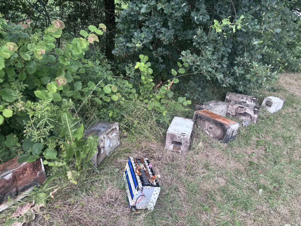 Mehrere verrostete Metallkästen sind auf einer Wiese abgelegt, teilweise im Gebüsch verborgen. Das Bild zeigt eine ländliche Gegend mit unordentlichem Abfall.
