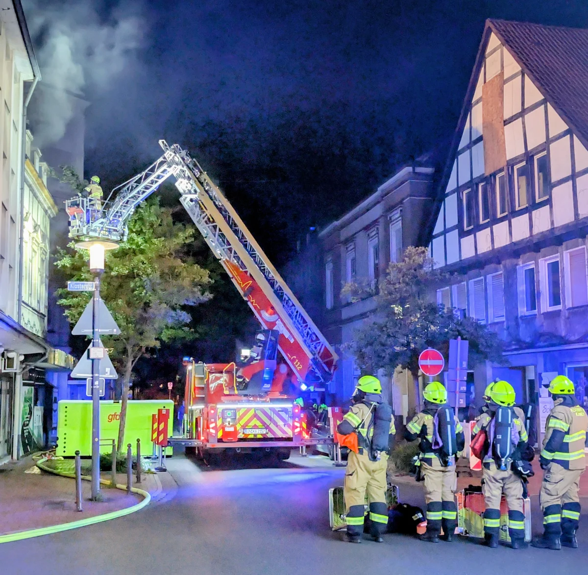 Ein nächtlicher Einsatz der Feuerwehr zeigt Einsatzkräfte und einen Leiterwagen vor einem rauchenden Gebäude in einer Stadt. Feuerwehrleute in Schutzanzügen beobachten die Lage.