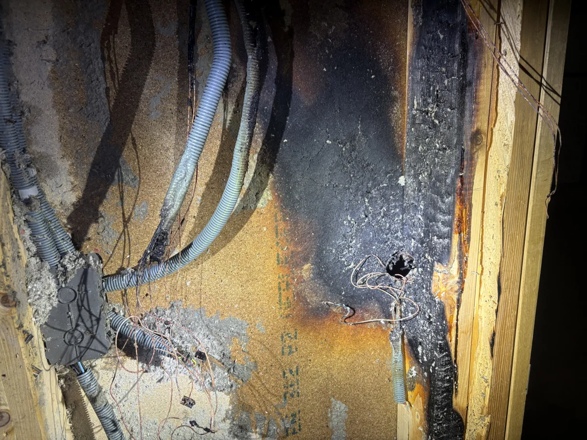 Die Wand zeigt deutliche Spuren eines Schmorbrandes mit stark beschädigten und verkohlten Kabeln. Der Putz ist teilweise abgebröckelt und die Verkohlung breitet sich über die Oberfläche aus.