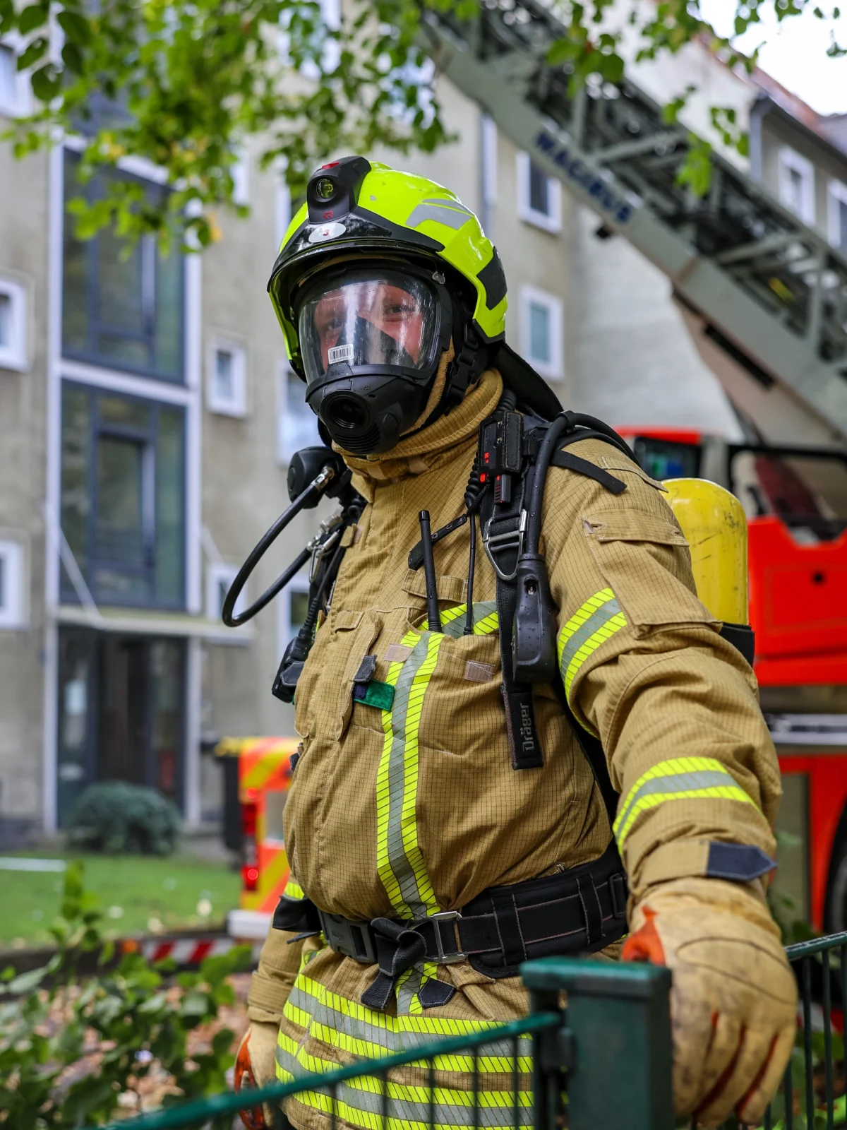 Ein Feuerwehrmann trägt Schutzkleidung, Atemmaske und Helm, steht vor einem Mehrfamilienhaus mit Einsatzfahrzeugen im Hintergrund.