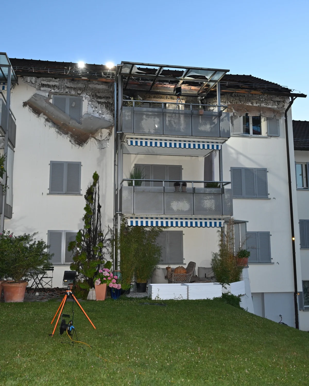 Das Bild zeigt die Fassade eines Wohnhauses mit sichtbaren Brandschäden am oberen Balkon und Dach. Im Vordergrund ist ein vermessenes Tripod auf einem Rasen zu sehen.