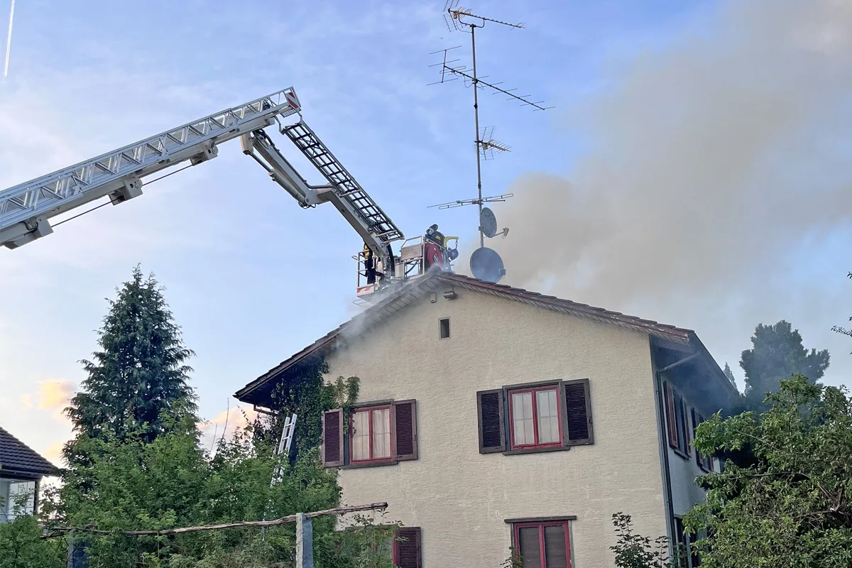 Ein Leiterwagen der Feuerwehr ist an einem Hausdach positioniert, wo Feuerwehrleute einen Brand löschen. Rauch steigt auf.