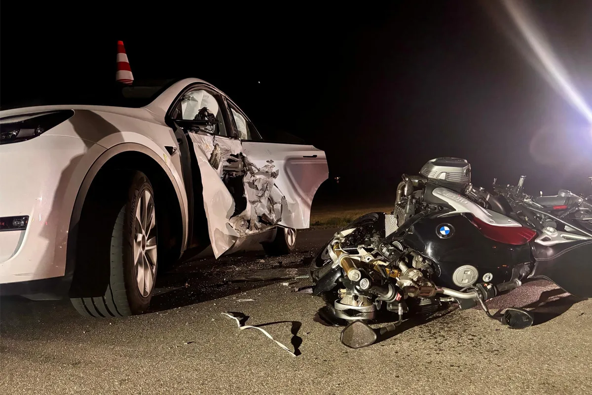 Ein weißes Auto mit schweren Schäden an der Seite steht neben einem am Boden liegenden BMW-Motorrad. Die Szene wird von einem Scheinwerferlicht beleuchtet.