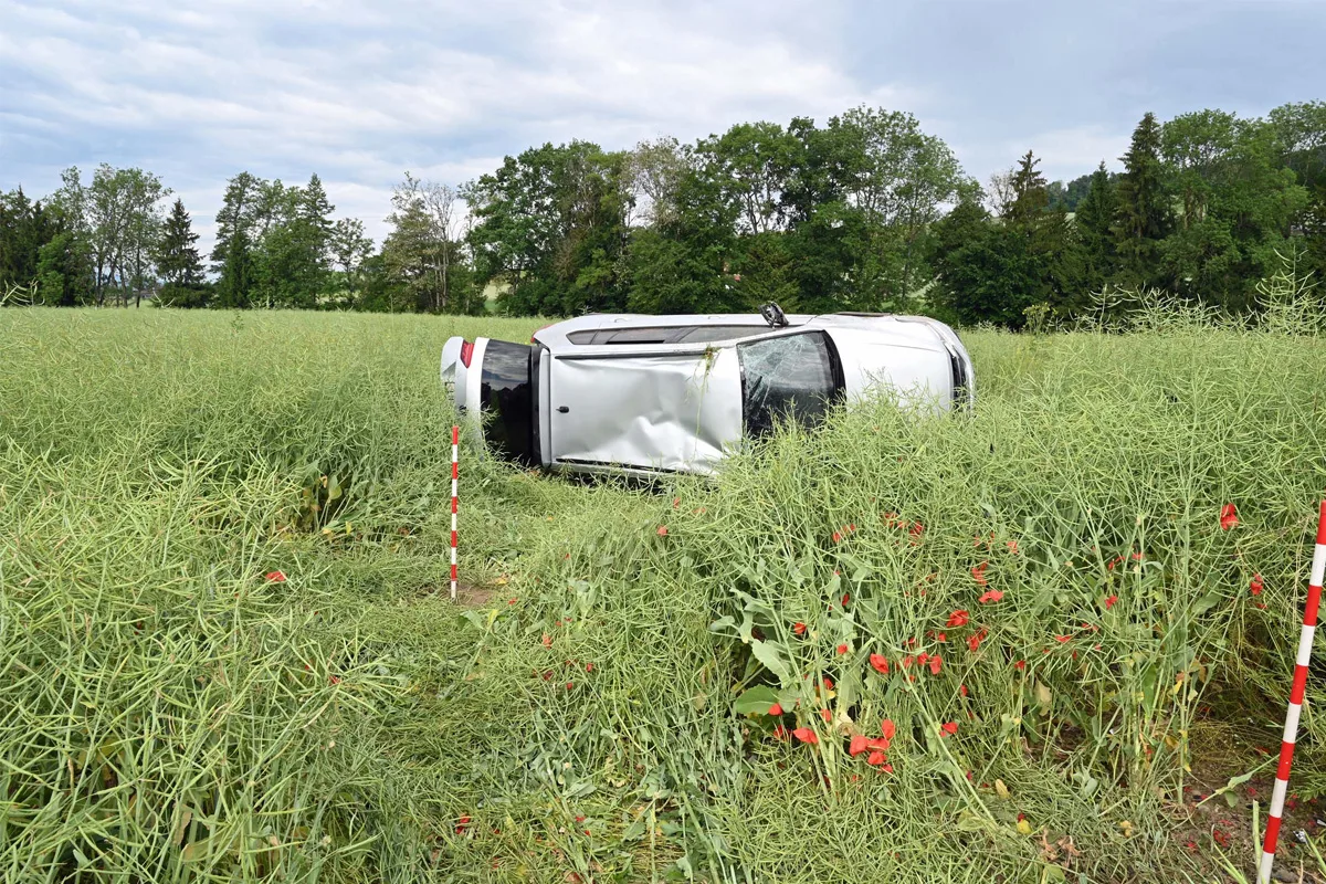 Ein weißes Auto liegt auf der Seite in einem Feld mit hohem Gras und roten Markierungsstäben vorn im Bild.