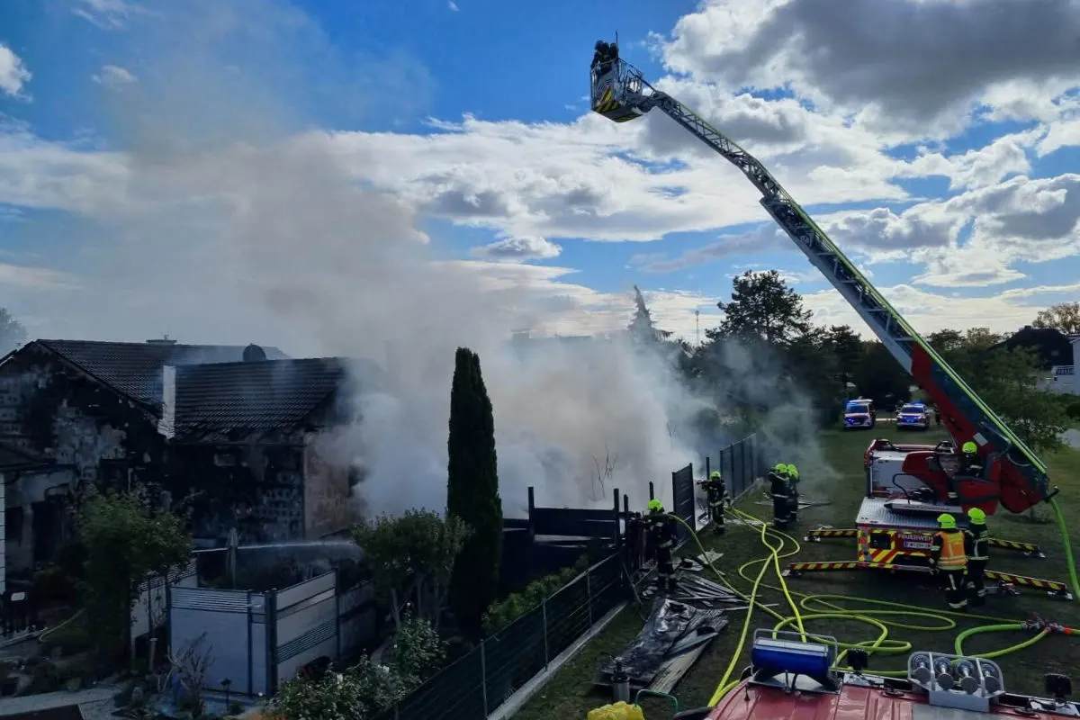 Ein großes Wohnhaus steht in Flammen, während Feuerwehrkräfte mit einem Leiterfahrzeug den Brand bekämpfen. Rauch steigt aus dem Gebäude auf, und Schläuche liegen am Boden.