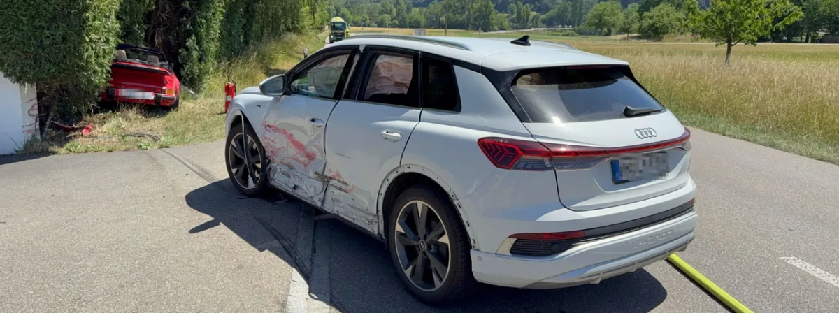 Ein weißer Audi mit Schäden an der Seite steht auf einer Landstraße. Im Hintergrund ist ein roter Sportwagen im Gras sichtbar, der ebenfalls verunfallt ist.