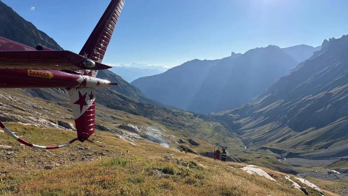 Ein rotes Helikopterheck ragt in eine weite Berglandschaft, die unter klarem, sonnigem Himmel liegt. Zwei Personen in Rettungsausrüstung stehen in der Entfernung auf der Wiese.