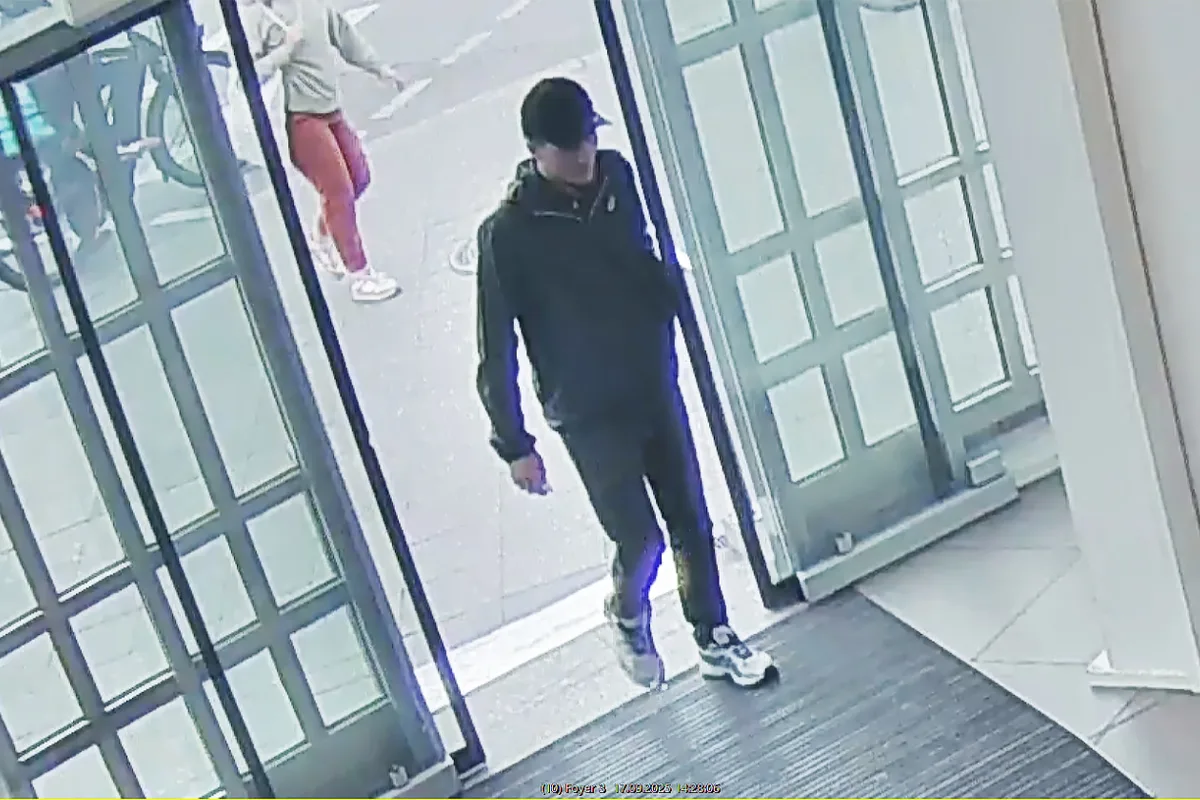 Ein Mann in dunkler Kleidung und Mütze betritt durch eine Glastür ein Gebäude. Im Hintergrund ist eine andere Person zu sehen.