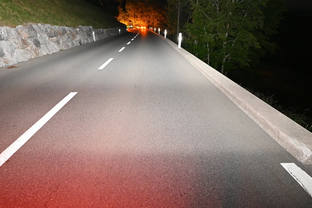Eine leere Landstraße, die bei Nacht von Scheinwerfern beleuchtet wird. Am Rand sind Bäume und Felsen sichtbar.