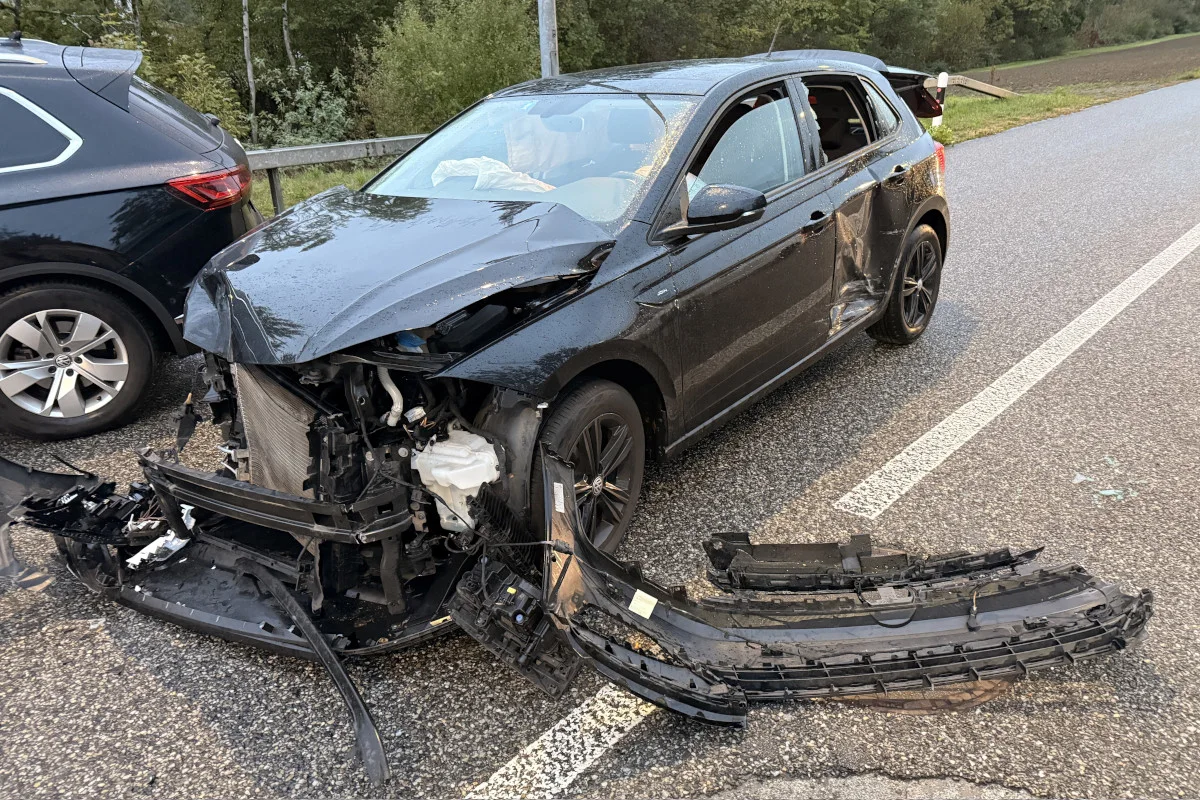 Ein schwarzes Auto ist stark beschädigt nach einem Unfall auf einer Straße. Der Frontbereich ist eingedrückt und der Stoßfänger liegt auf dem Boden.