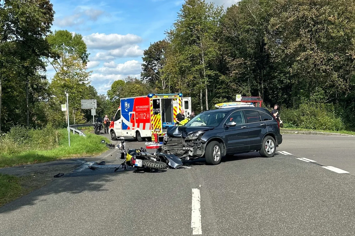 Ein schwarzes Auto mit stark beschädigter Front steht an einer Kreuzung, neben einem umgestürzten Motorrad. Ein Rettungswagen ist vor Ort.