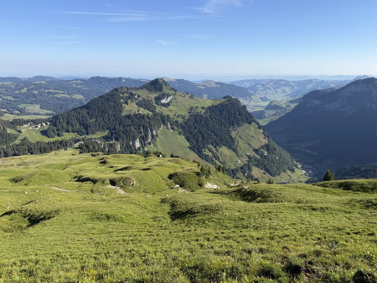 Eine weite grüne Berglandschaft mit sichtbaren Tälern und Hügeln erstreckt sich bis zum Horizont. Der Himmel ist klar und blau.