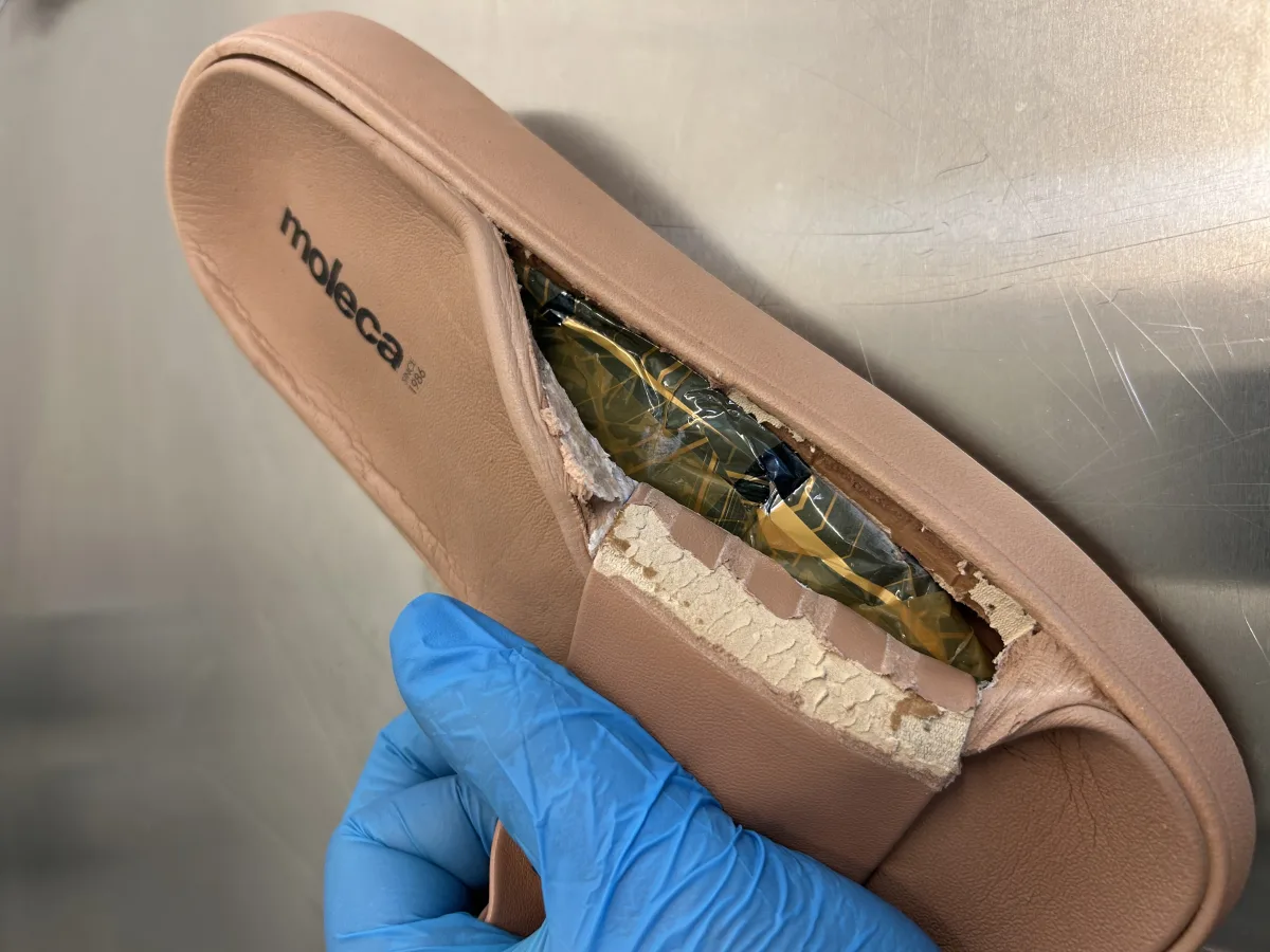 Eine Hand in einem blauen Handschuh hält einen aufgeschnittenen Schuh, der ein verstecktes Drogenpaket zeigt. Der Schuh hat eine beige Farbe, und das Paket ist in einer glänzenden Verpackung.