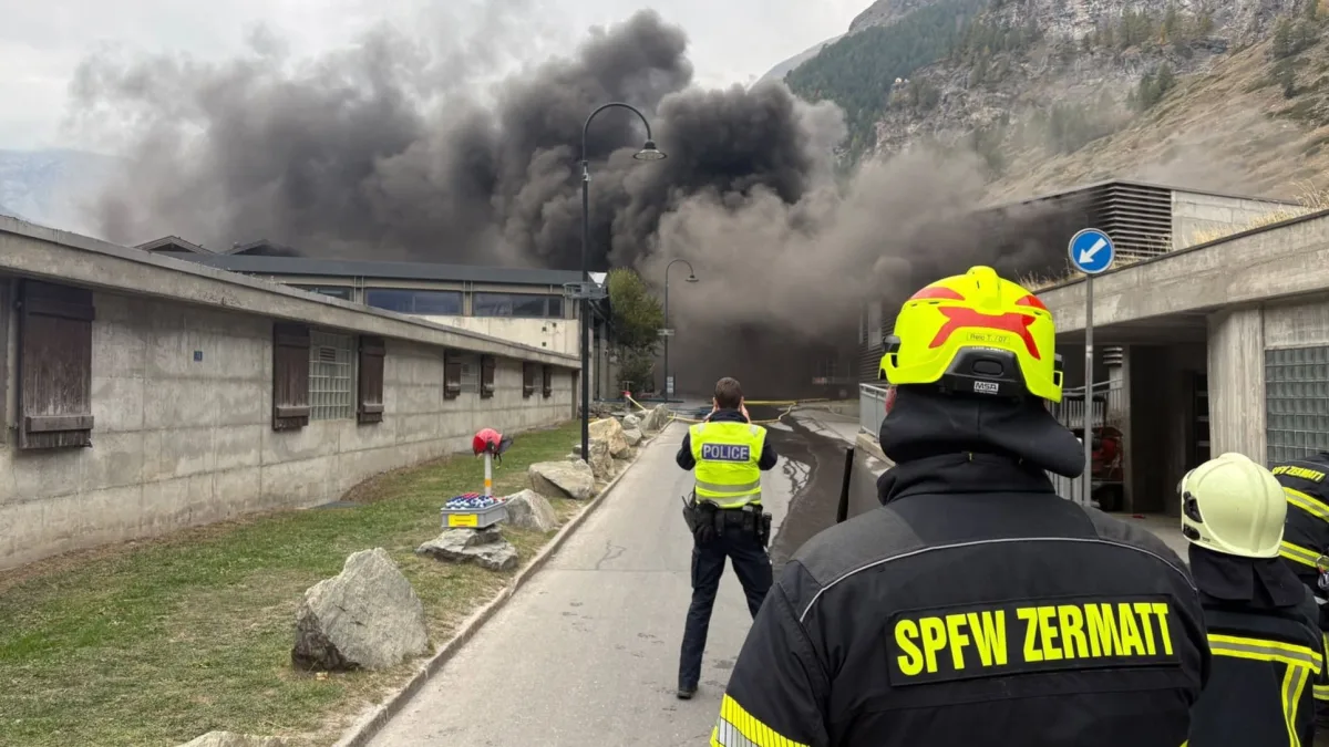 Eine massive Rauchwolke erhebt sich aus einem Gebäude. Im Vordergrund befinden sich Feuerwehr- und Polizeikräfte, die die Lage überwachen.