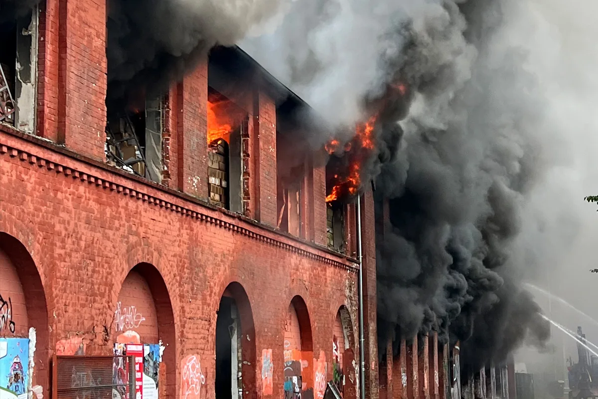 Ein großes verlassenes Backsteingebäude steht in Flammen. Dichte schwarze Rauchwolken steigen aus den oberen Fenstern auf, während Feuer im Inneren lodert.