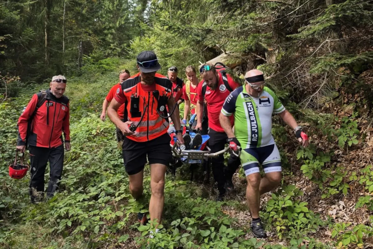 Mehrere Personen, darunter Rettungskräfte, tragen eine verletzte Person auf einer Trage durch dicht bewaldetes Terrain. Sie tragen rote Rettungswesten.