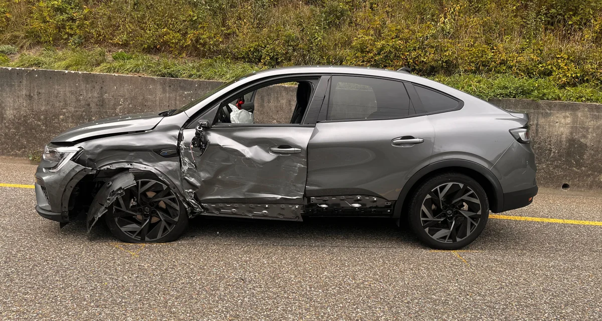 Ein silbernes Auto zeigt schwere Beschädigungen an der Seite; es parkt am Rand einer Straße mit grüner Böschung im Hintergrund.
