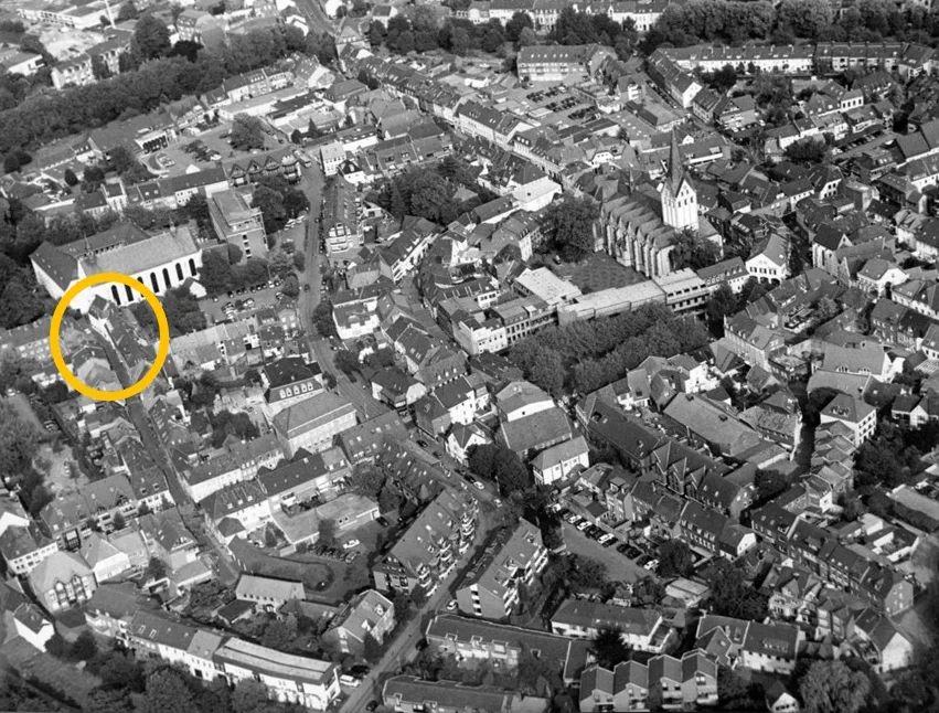 Die Schwarz-Weiß-Luftaufnahme zeigt eine Stadt mit dicht bebauten Strukturen. Ein gelber Kreis hebt ein Gebäude hervor.