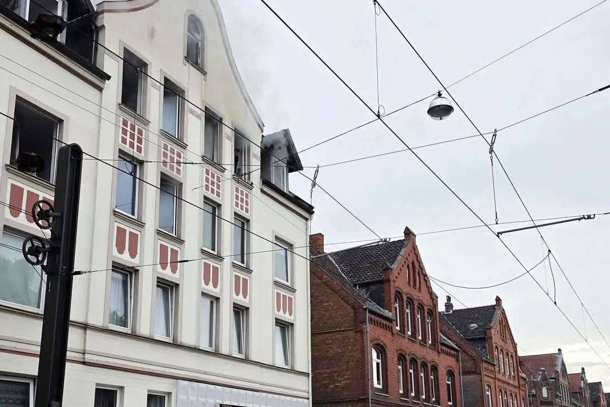 Ein mehrstöckiges Gebäude mit Rauch, der aus einem Dachfenster austritt. Mehrere Nachbargebäude sind sichtbar.