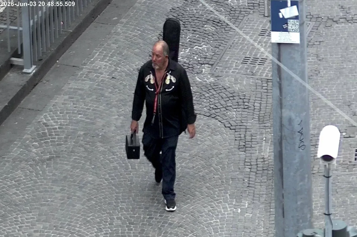 Ein älterer Mann trägt einen kleinen Koffer und geht auf einer gepflasterten Straße, aufgenommen von einer Überwachungskamera bei Tageslicht.