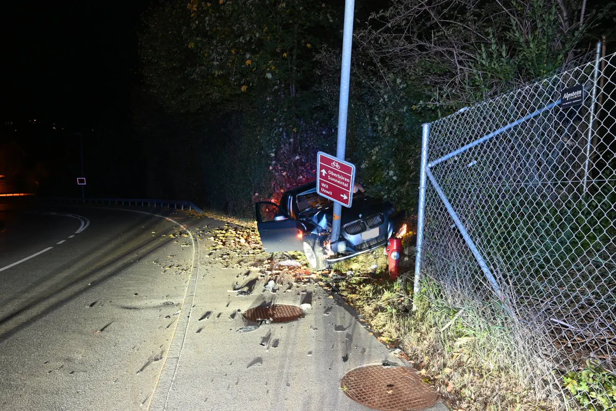 Ein Auto mit geöffneten Türen kollidiert mit einem Laternenmast und einem Verkehrsschild an einer kurvigen Straße. Trümmerteile sind auf dem Boden verstreut.