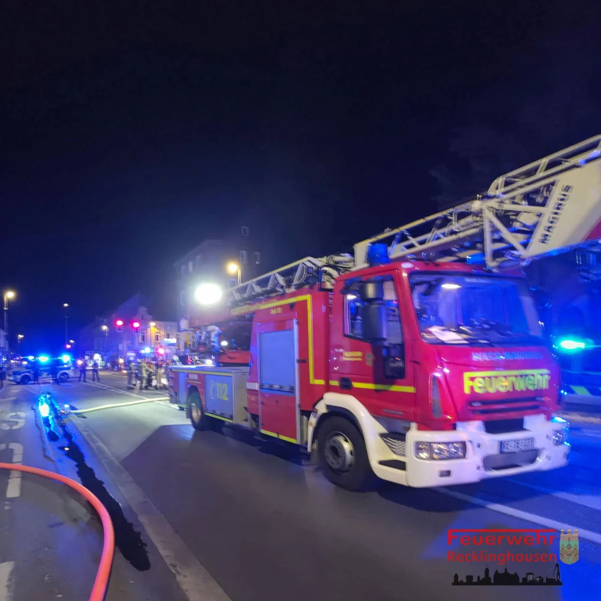 Ein rotes Feuerwehrfahrzeug mit Leiter steht nachts auf einer Straße, umgeben von blauem Einsatzlicht und mehreren Personen.
