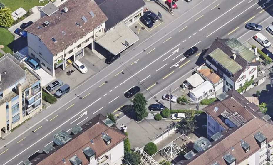 Die Luftaufnahme zeigt eine Kreuzung mit mehreren Fahrspuren, umgeben von Wohngebäuden. Geparkte Autos sind entlang der Straße zu sehen.