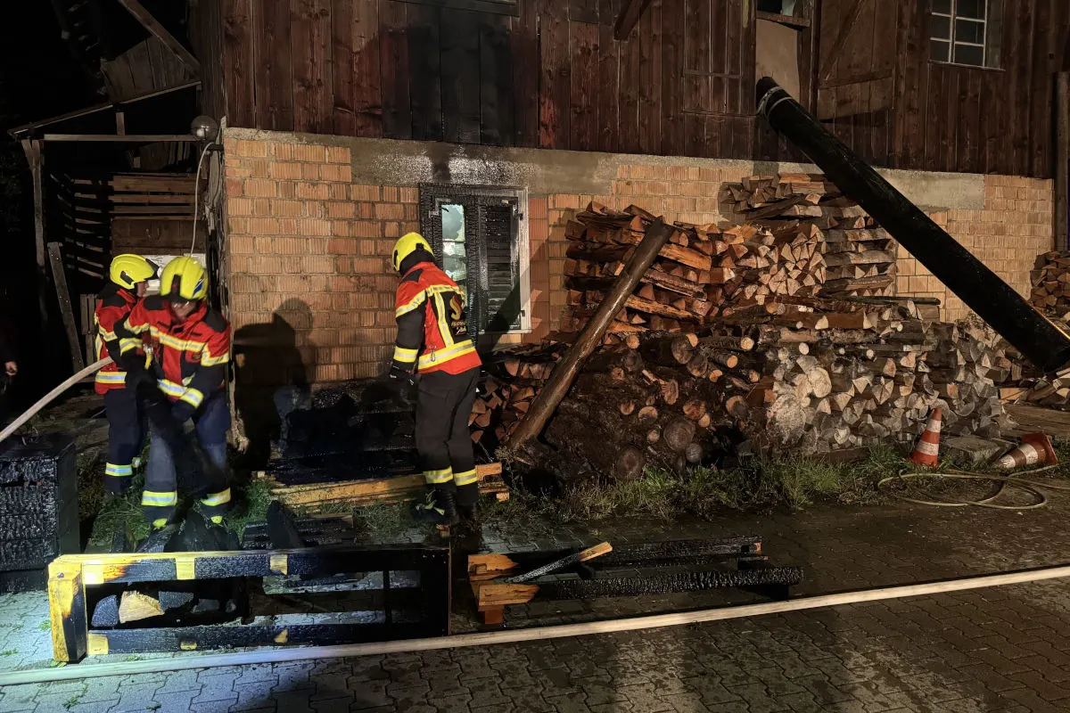 Drei Feuerwehrleute in Schutzkleidung löschen mit einem Schlauch ein Feuer an der Seite eines Gebäudes. Im Hintergrund sind gestapelte Holzscheite sichtbar.