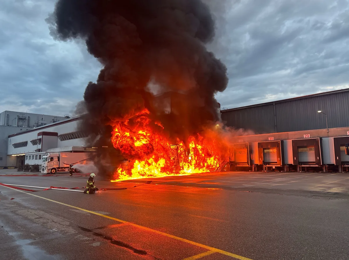 In einem Gewerbegebiet steht ein Gebäude in Flammen. Dichter schwarzer Rauch steigt auf, während Feuerwehrleute das Feuer bekämpfen.