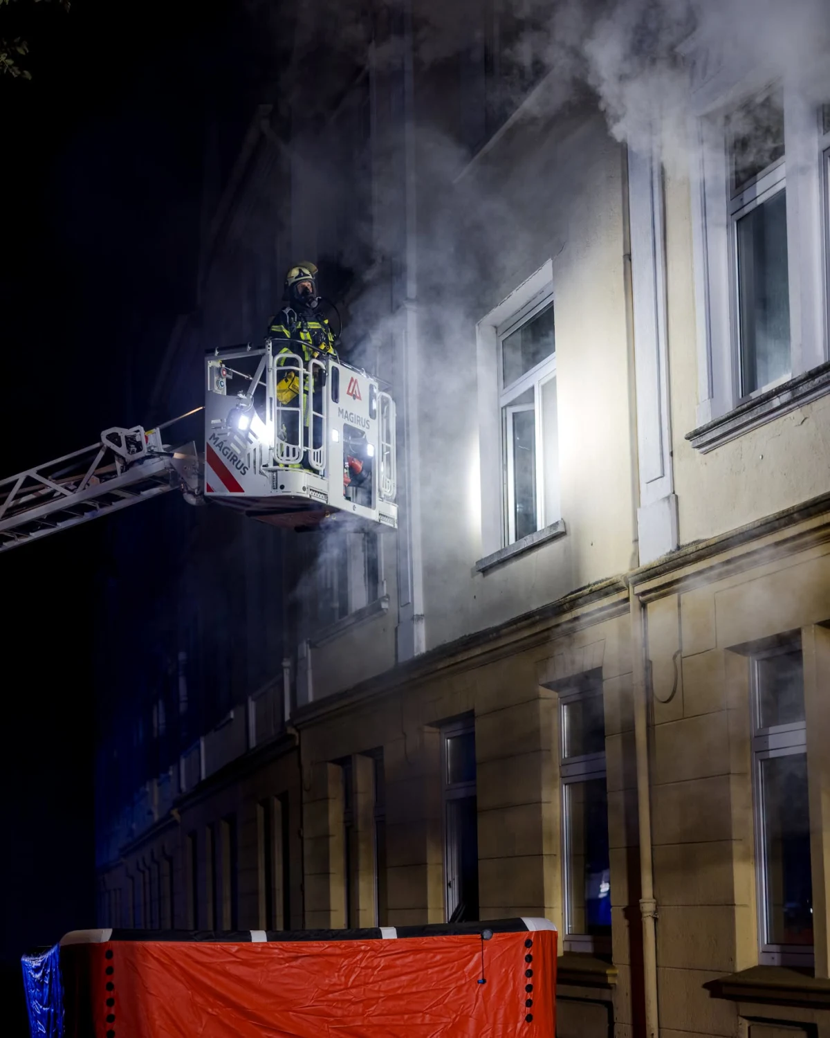Ein Feuerwehrmann in Schutzkleidung steht auf einer Drehleiter vor einem Gebäude, aus dem Rauch tritt. Das Szenario spielt sich nachts ab.