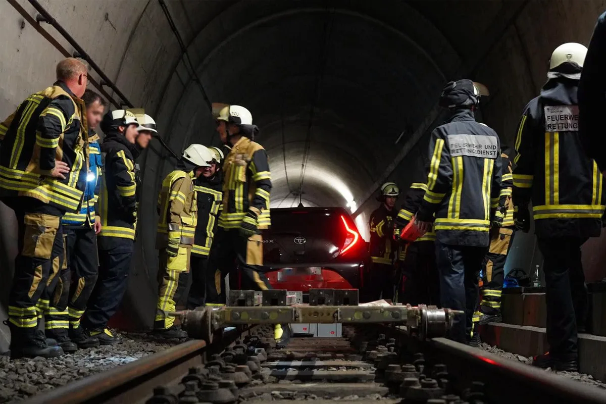 Feuerwehrleute stehen um ein schwarzes Auto herum, das in einem Eisenbahntunnel feststeckt. Der Tunnel ist dunkel und die Szene wird von den reflektierenden Streifen der Uniformen erhellt.