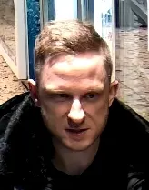 Ein Mann mit kurzen Haaren und ernstem Gesichtsausdruck befindet sich in einem Innenraum mit Glasflächen.