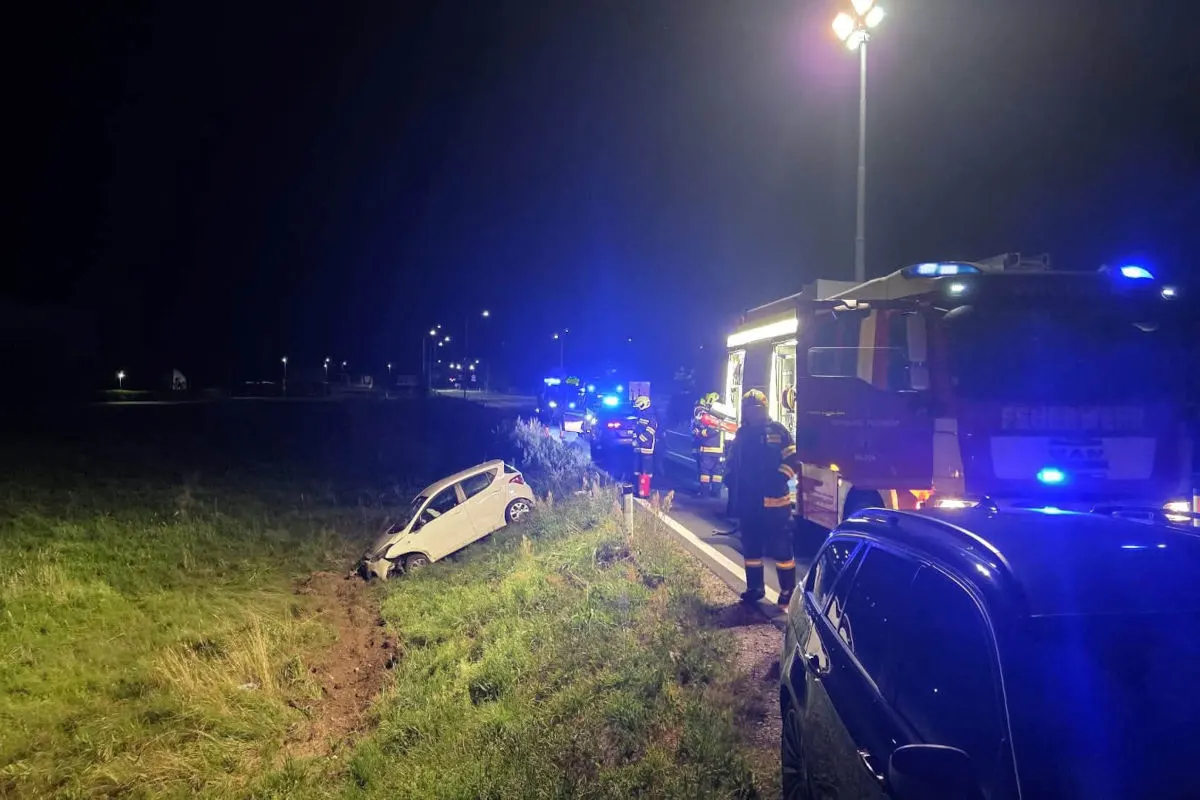 Ein weißes Auto liegt in einem Graben, während Feuerwehrfahrzeuge und Einsatzkräfte bei Nacht auf der Straße zu sehen sind.