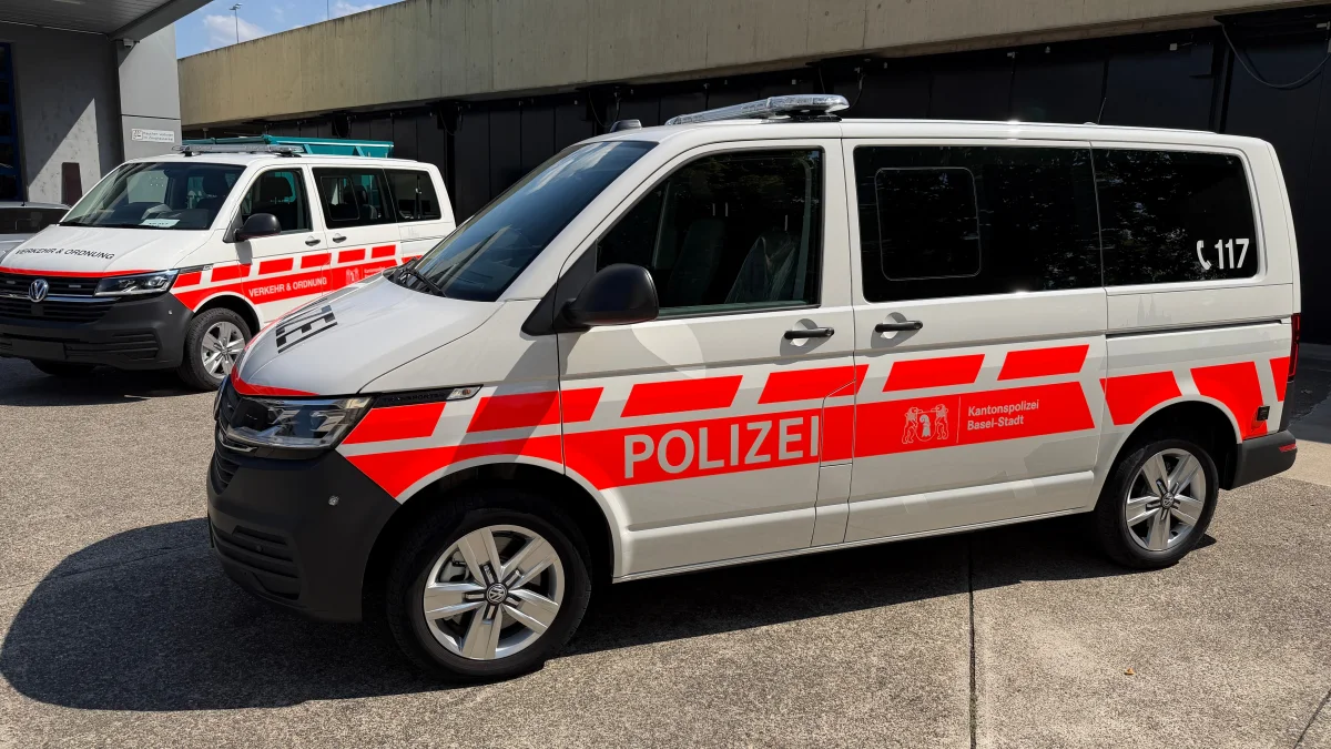 Zwei weiße VW-Transporter mit roten Streifen und der Aufschrift 'Polizei' der Kantonspolizei Basel-Stadt stehen im Freien.