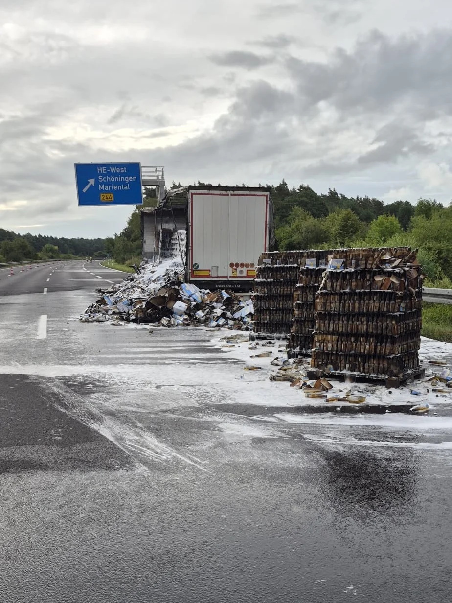 Ein stark beschädigter Lkw-Anhänger mit verbrannter Ladung steht am Straßenrand. Daneben liegen Getränkekisten auf der Fahrbahn.