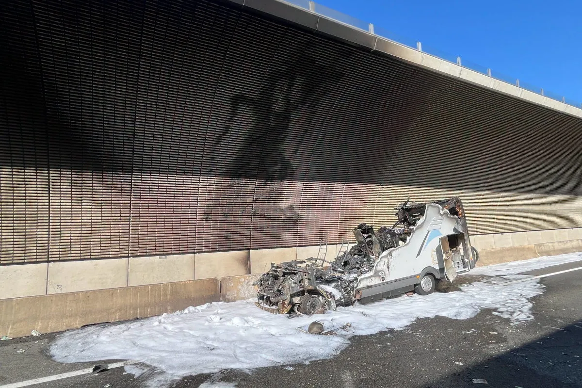 Ein stark beschädigtes, ausgebranntes Wohnmobil liegt vor einer Tunnelwand. Auf der Straße ist Löschschaum zu sehen.