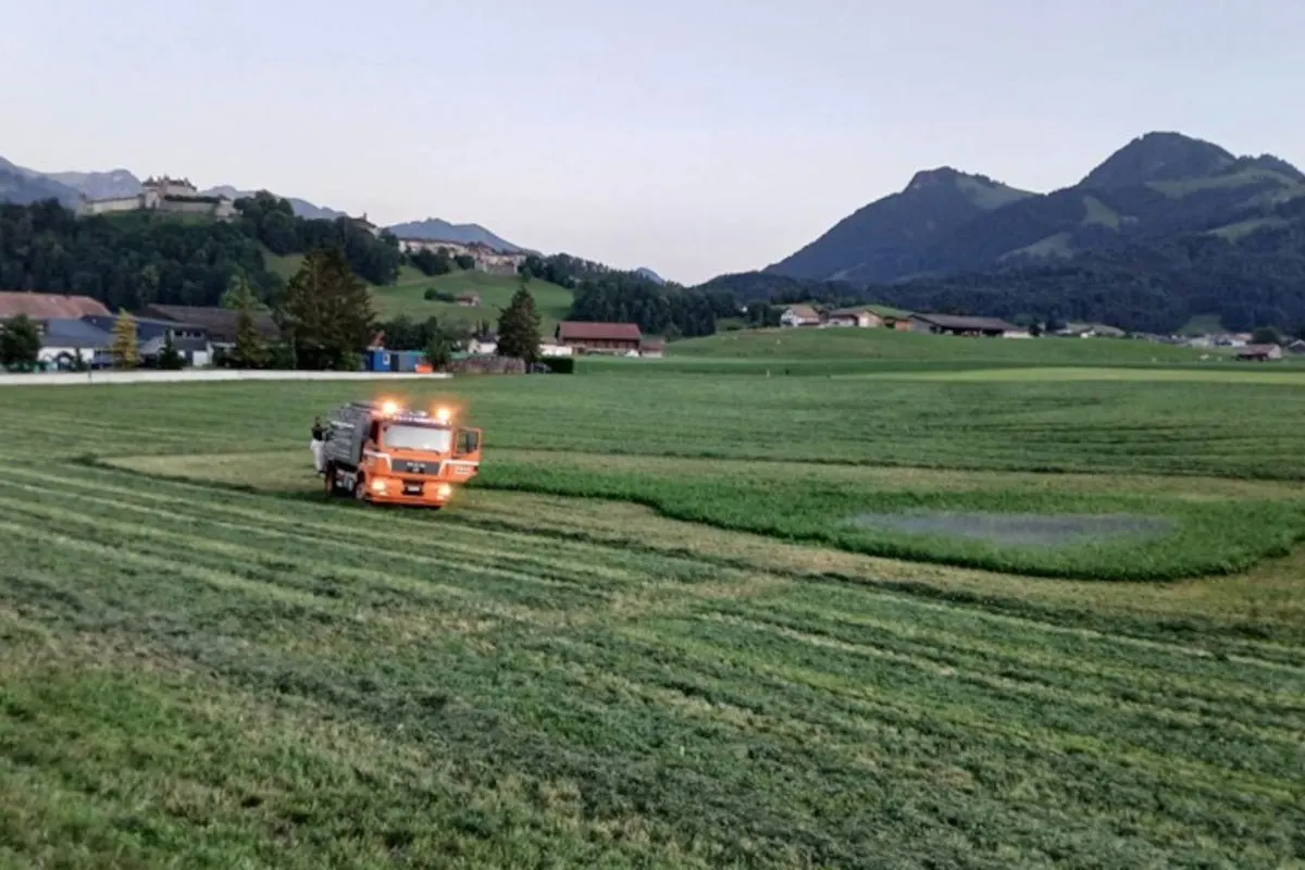 Ein orangefarbener Lastwagen mit Blinklicht steht auf einer großen, grünen Wiese. Im Hintergrund sind Gebäude und Berge zu sehen.